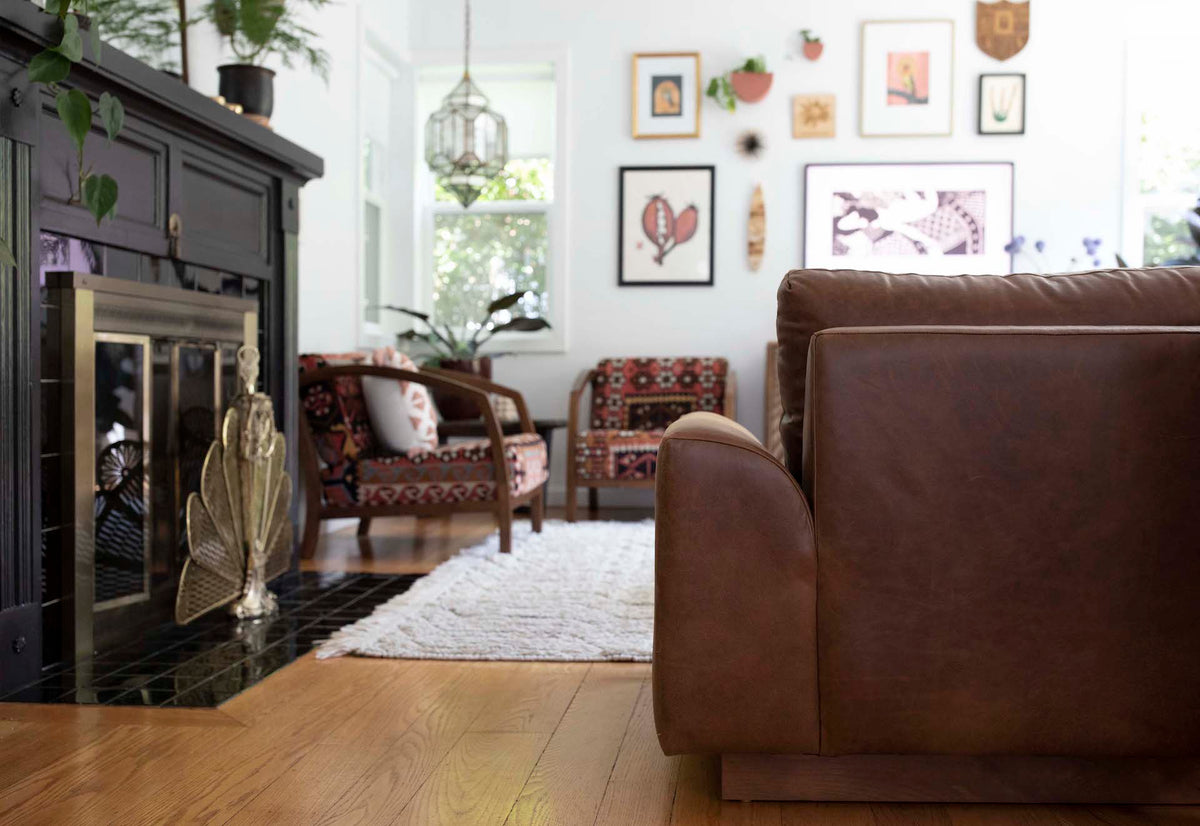 Back of a brown leather couch in a modern living room with chairs, a rug, a black fireplace, and different art prints hung on the wall.