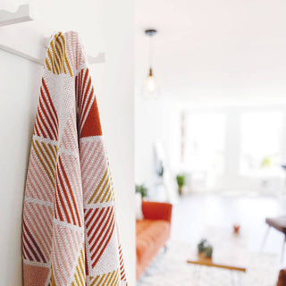 A Seek and Swoon Zion Throw, which features a geometric design with orange, gold and blush striped triangles hanging on a hook in a bright, modern room with an orange couch just visible in the background. 