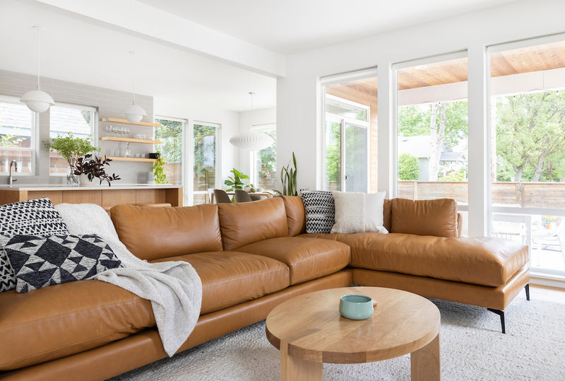 Modern living room with a brown leather sectional sofa, wooden coffee table, and large windows.