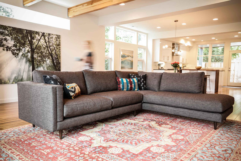 grey sectional couch in a living room, with a patterned rug and decorative pillows on top and blurry movement of a person behind the sectional.
