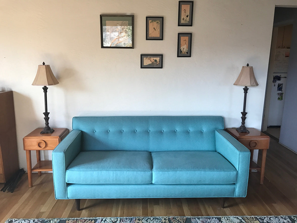 A turquoise couch with two wooden side tables on each side, with a lamp placed on each table and small prints hanging from the wall behind the couch.